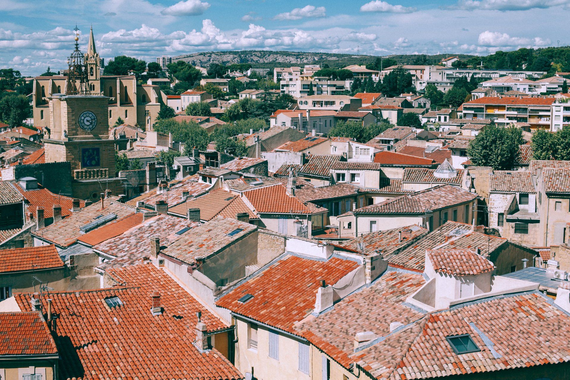 Vista de edificios con tejados de tejas rojizas, chimeneas y ventanas de cubierta, una torre con reloj, árboles y colinas bajo un cielo con nubes.